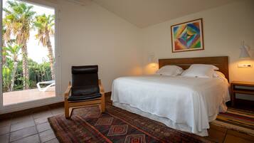 A hotel room with a bed, a chair, a colorful painting, and a view of palm trees through a glass door.