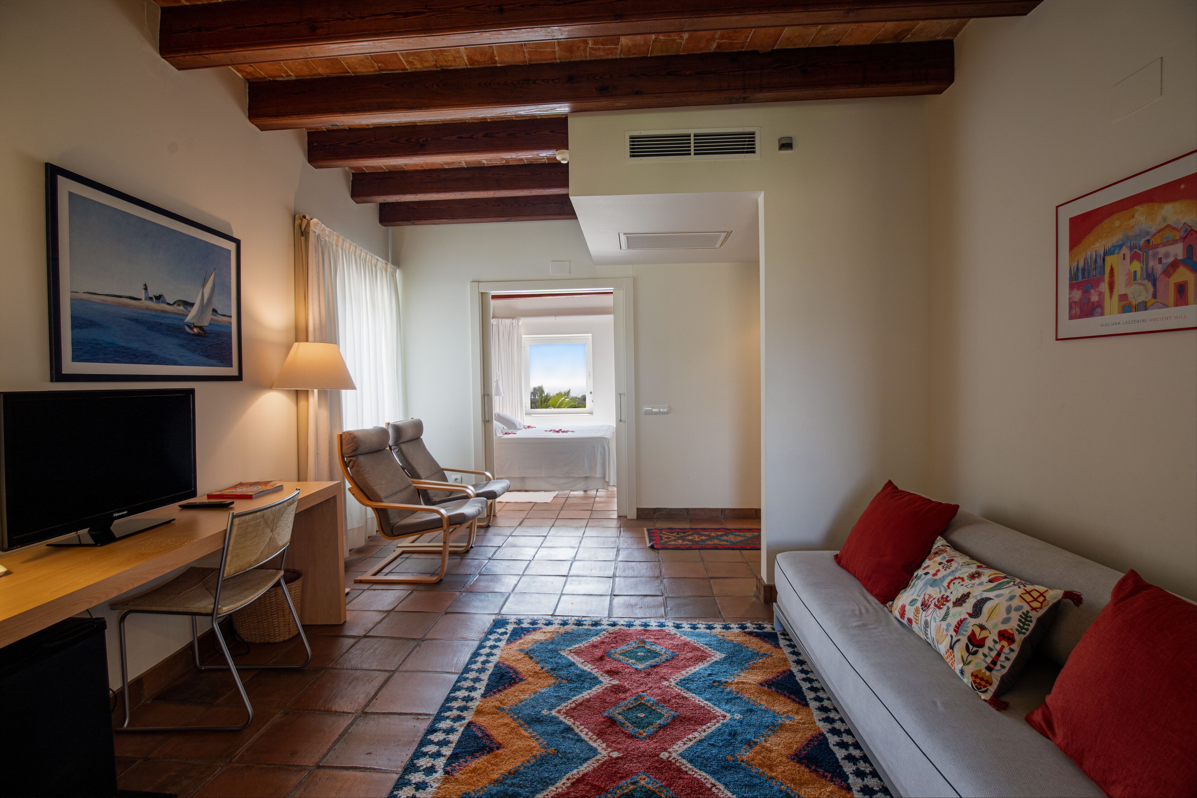 A living room with a sofa, a TV, a desk, and a chair. There is a window with curtains, a door leading to another room, and a colorful rug on the floor.