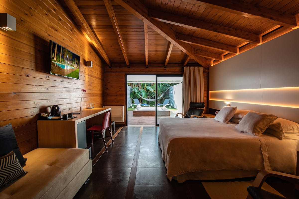 Habitación de hotel moderna con techo de madera, una cama grande, un escritorio con silla y vistas a una piscina y follaje exterior.
