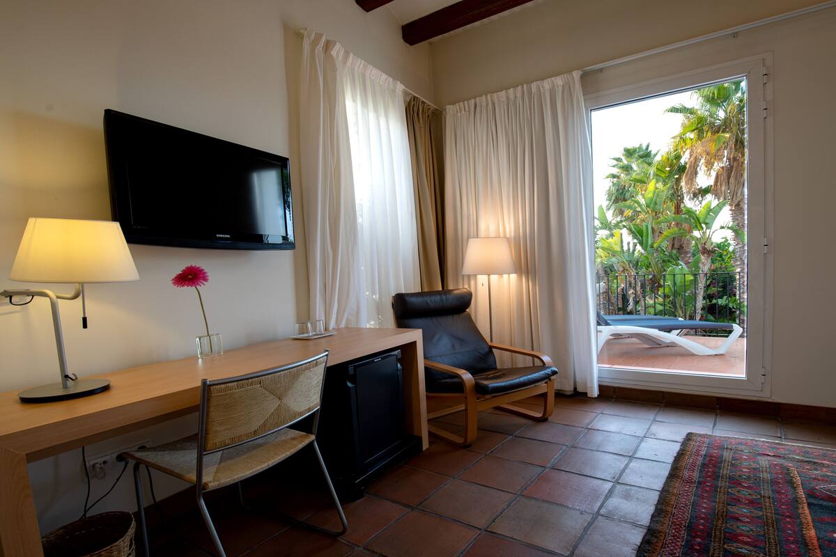 Habitación de hotel con televisor de pantalla plana, escritorio con lámpara, una silla, refrigerador pequeño y vista a un balcón con plantas.