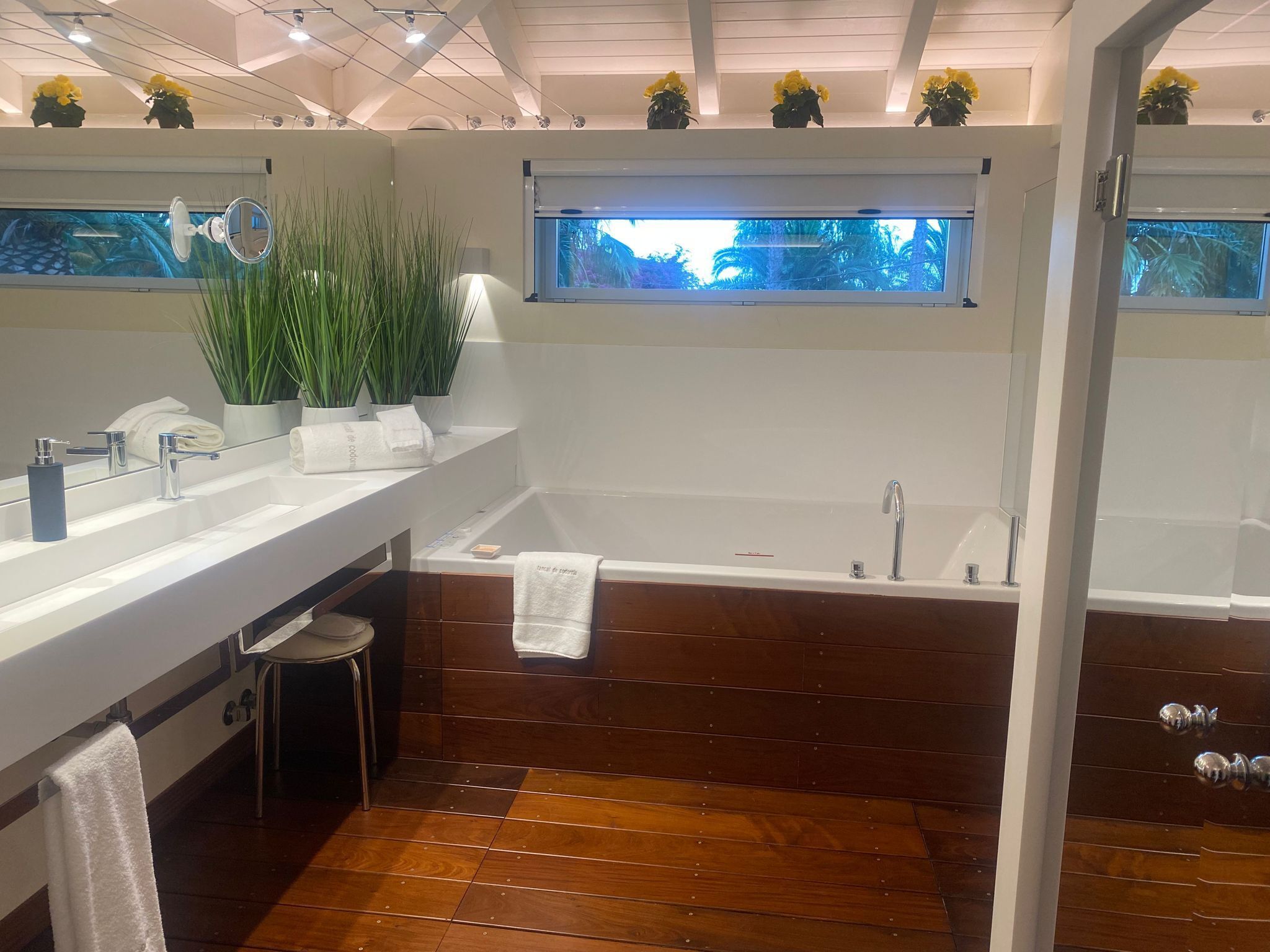 A modern bathroom with a wooden floor, a large bathtub, a white sink, and a window with a view of trees.