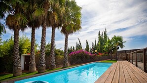 A swimming pool with wooden deck, surrounded by palm trees and a stone wall with flowering plants.