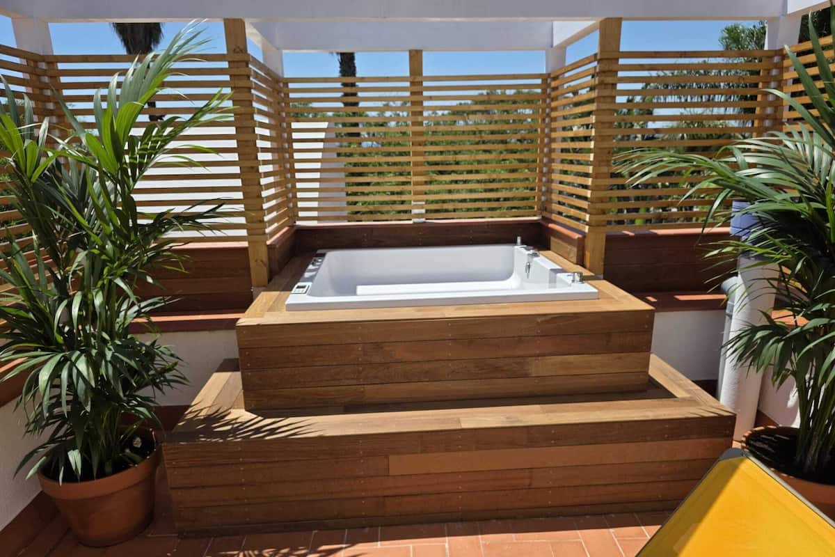 Un jacuzzi exterior de madera con escalones, rodeado de paneles de madera celosía y plantas en macetas.