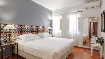 A hotel room with a large bed, two bedside tables with lamps, a colorful quilt on the headboard, a mirror, and a view of the hallway.