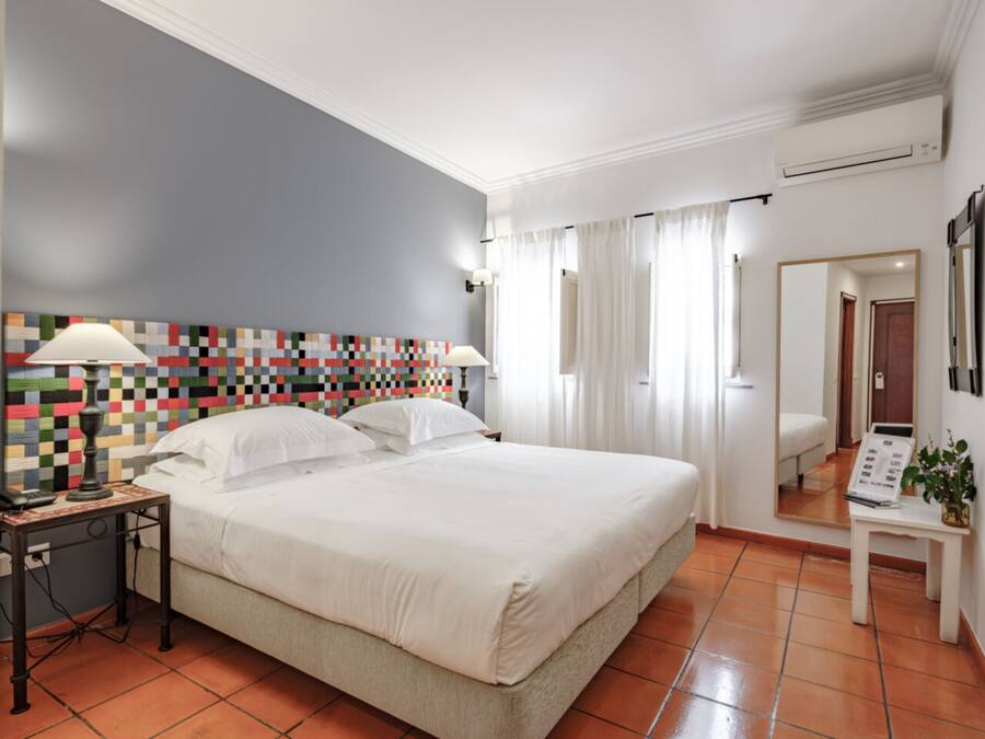 A hotel room with a large bed, two bedside tables with lamps, a colorful quilt on the headboard, a mirror, and a view of the hallway.