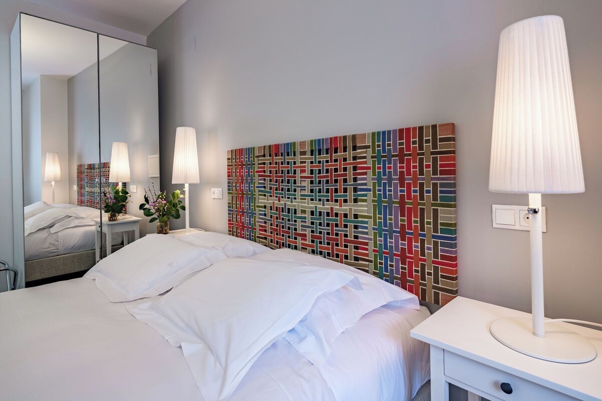 A modern hotel room with a large bed, a bedside table with a lamp, a mirror, and a colorful abstract wall art piece.