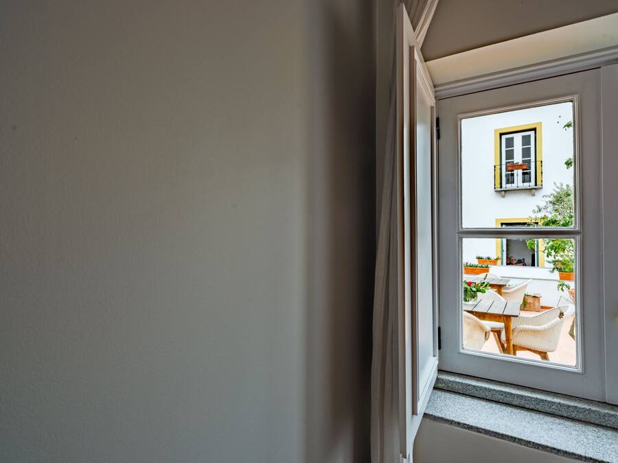 Uitzicht door een raam met wit frame, waarop een terras met tafels en stoelen en een gebouw met een geel raamkozijn te zien is.