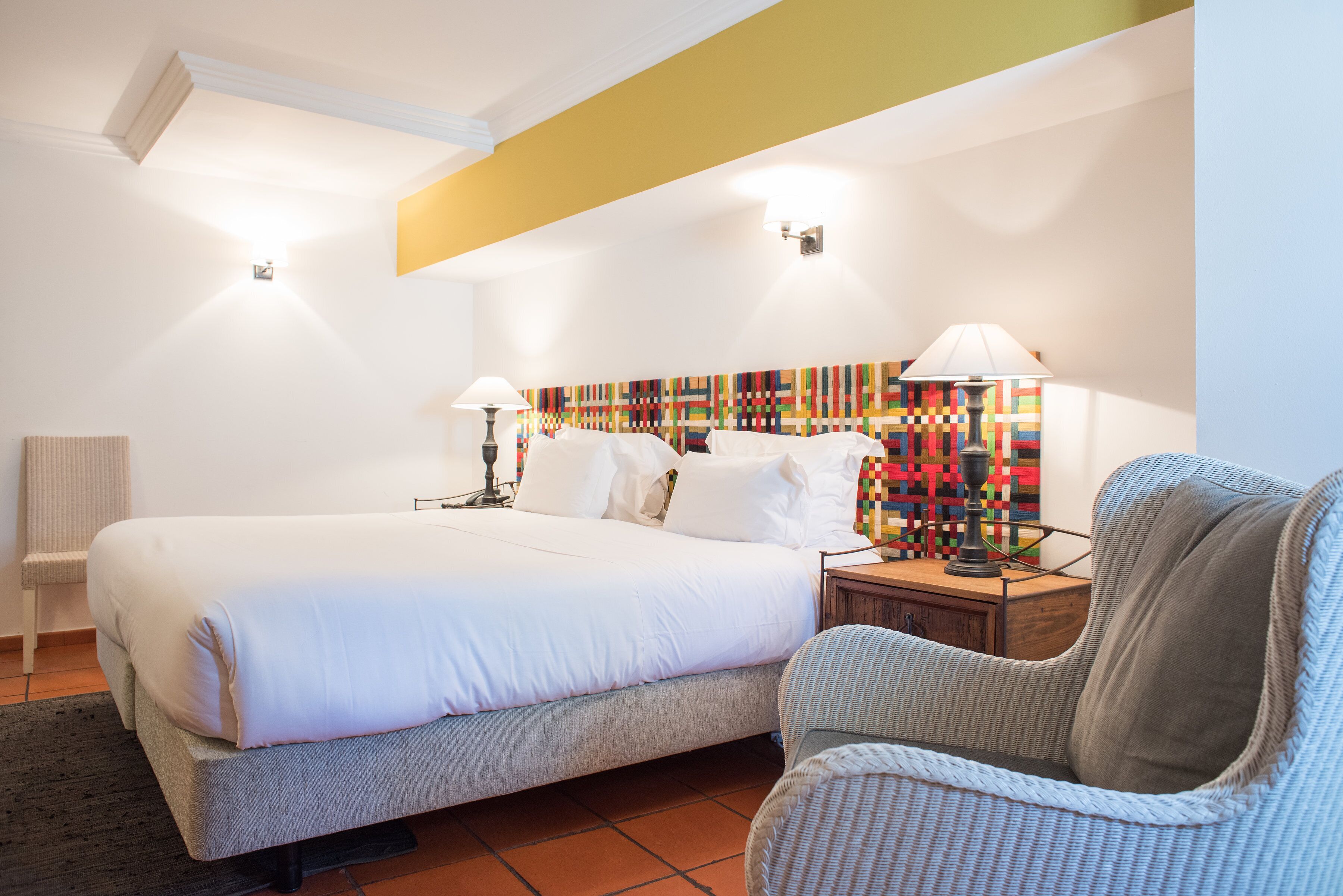 Une chambre d’hôtel avec un grand lit, deux lampes de chevet, une décoration murale colorée et un fauteuil en osier.