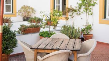 Un patio al aire libre con una mesa de madera y sillas de mimbre, rodeado de plantas en macetas y una pared blanca con detalles amarillos.