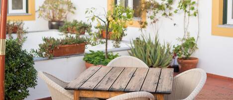 An outdoor patio with a wooden table and wicker chairs, surrounded by potted plants and a white wall with yellow trim.