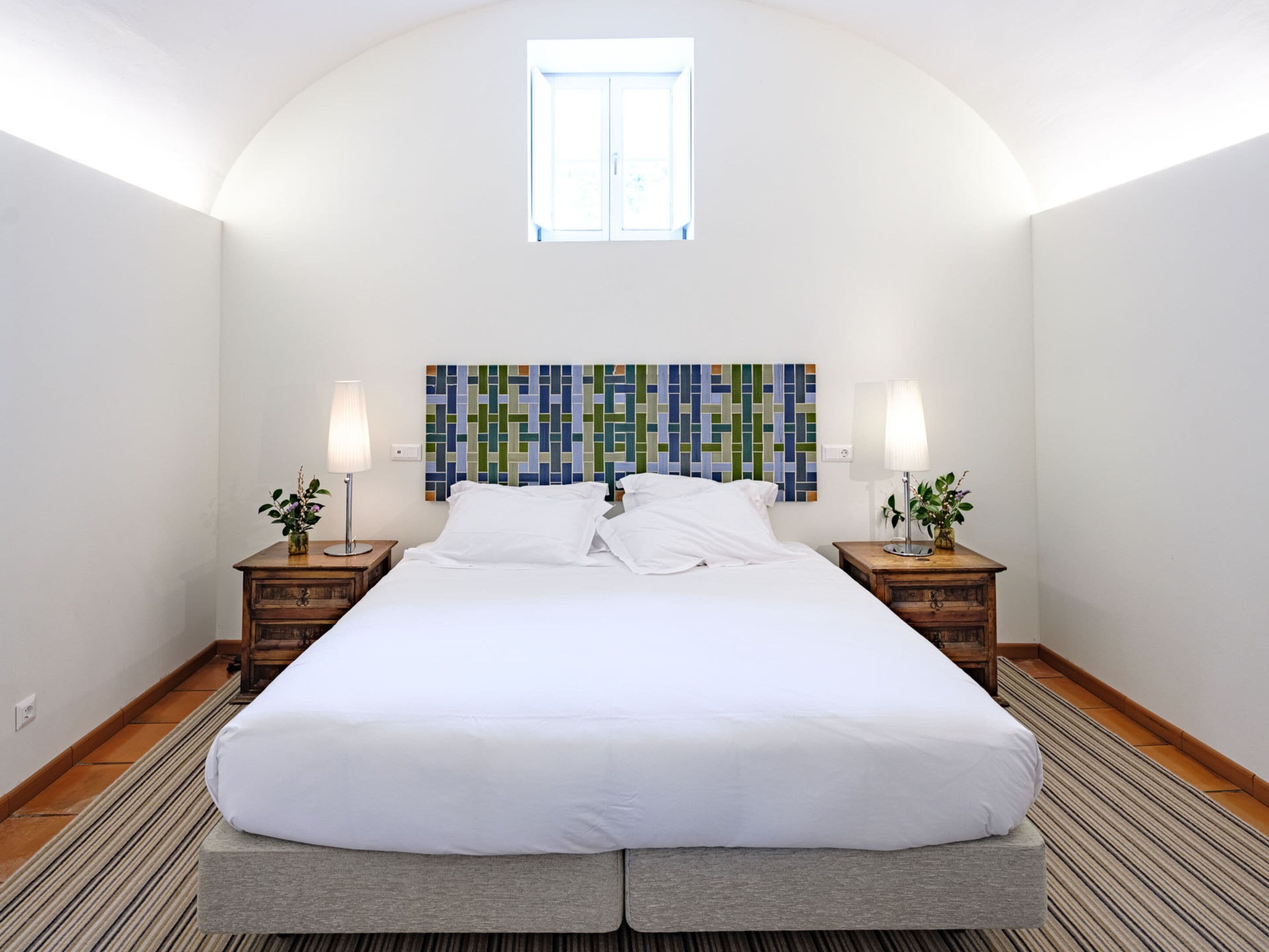 Una cama grande con ropa de cama blanca en una habitación con techo alto y abovedado y una ventana. Hay dos mesitas de noche con lámparas y un colorido tapiz abstracto sobre la cama.