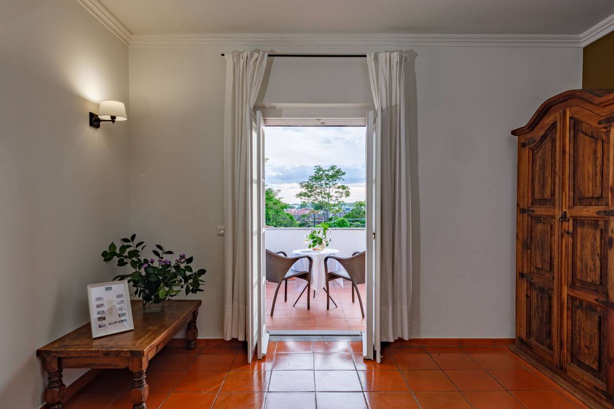 Una habitación con un armario de madera, un banco con una foto enmarcada, una planta en maceta y vistas a una zona de estar exterior a través de un vano.