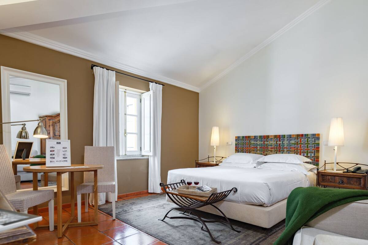 Una habitación de hotel con cama, mesitas de noche, escritorio, silla, ventana con cortinas y una obra de arte de colores en la pared.