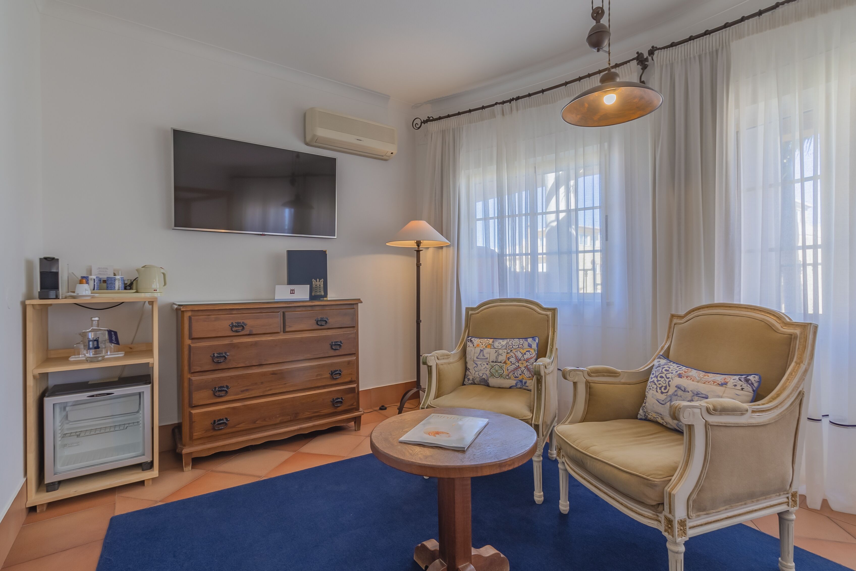 A room with a TV, wooden dresser, two chairs, a table, and a lamp.