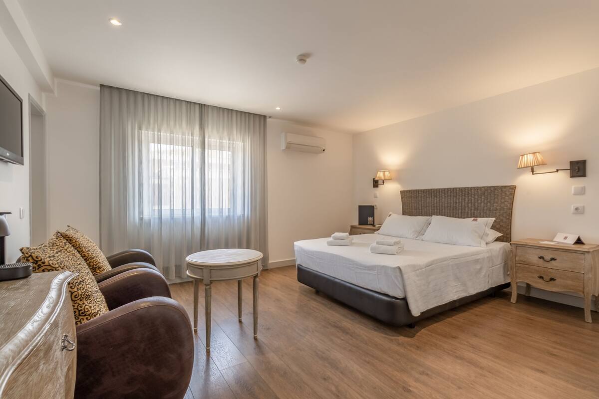 A modern hotel room with a large bed, a desk, and a chair. There is a window with curtains, a wall-mounted air conditioner, and a bedside lamp.