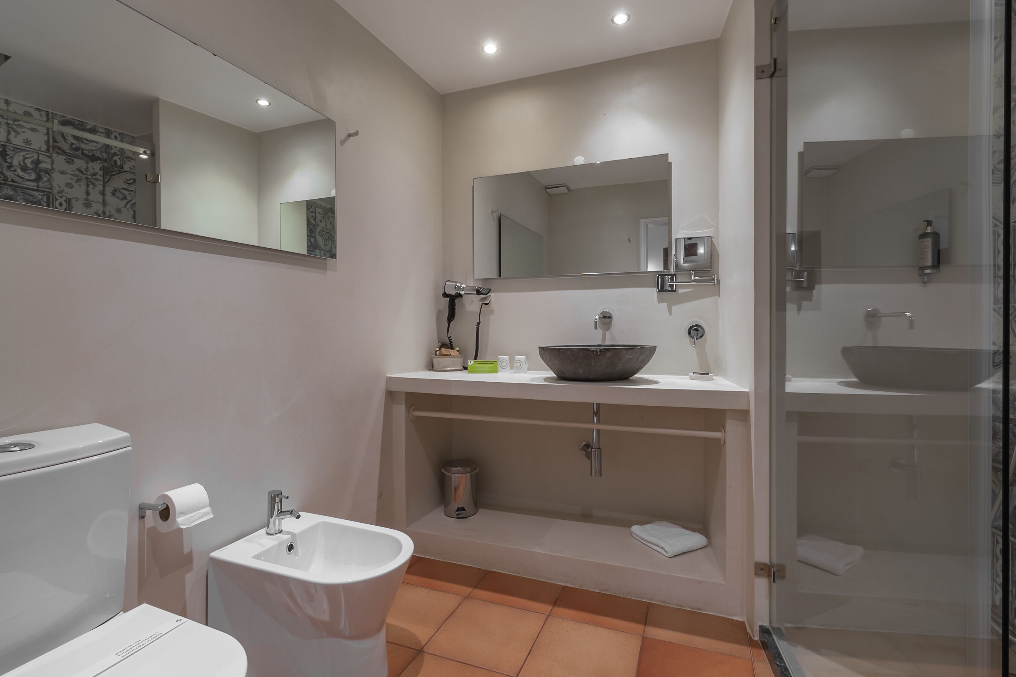 Ein modernes Badezimmer mit Toilette, Waschbecken und Bidet. Über dem Waschbecken hängt ein großer Spiegel, und die Dusche ist in eine Glasabtrennung eingebaut.