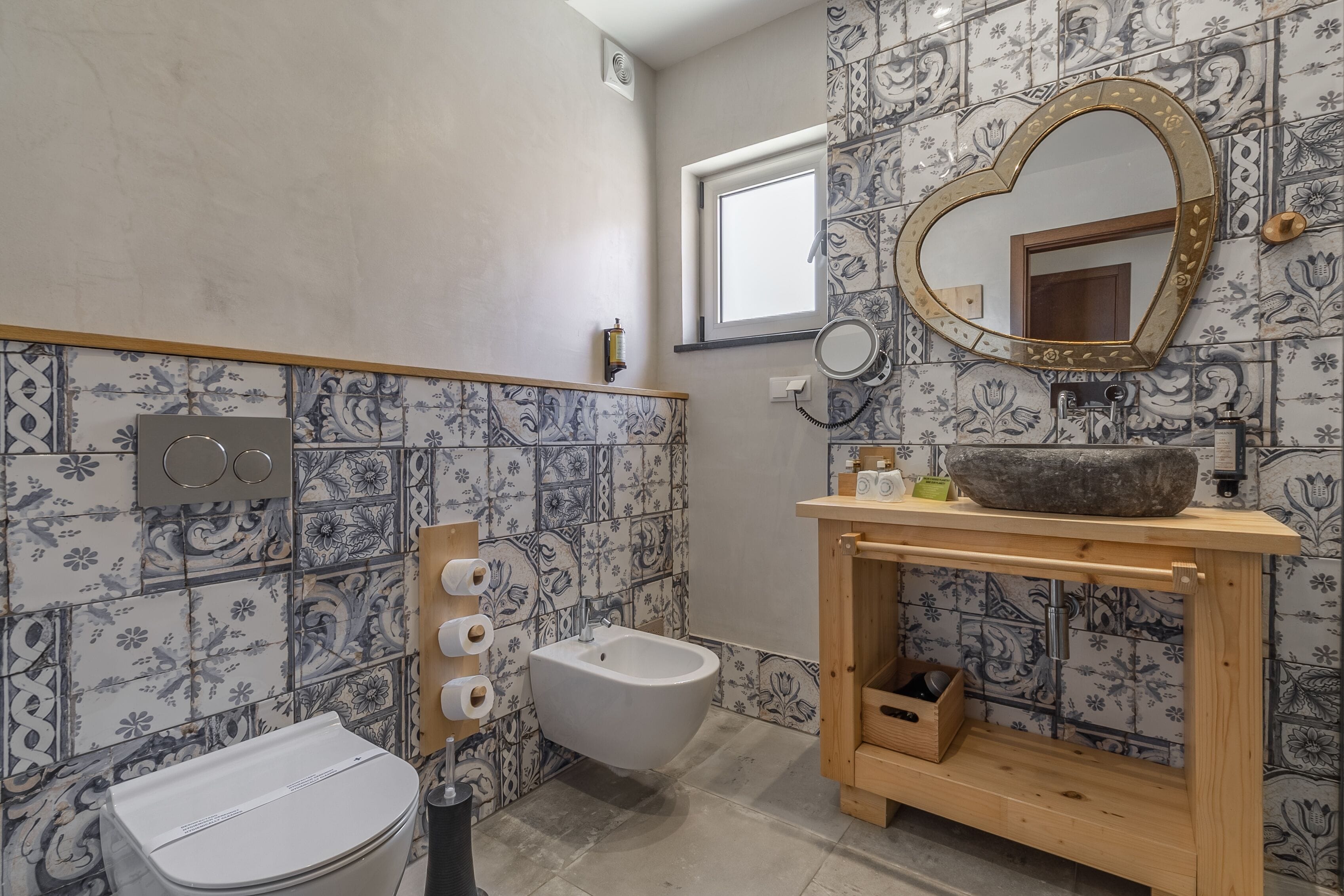 Une salle de bain équipée d’un bidet, de toilettes, d’un meuble-lavabo en bois avec un vasque, et d’un miroir en forme de cœur.