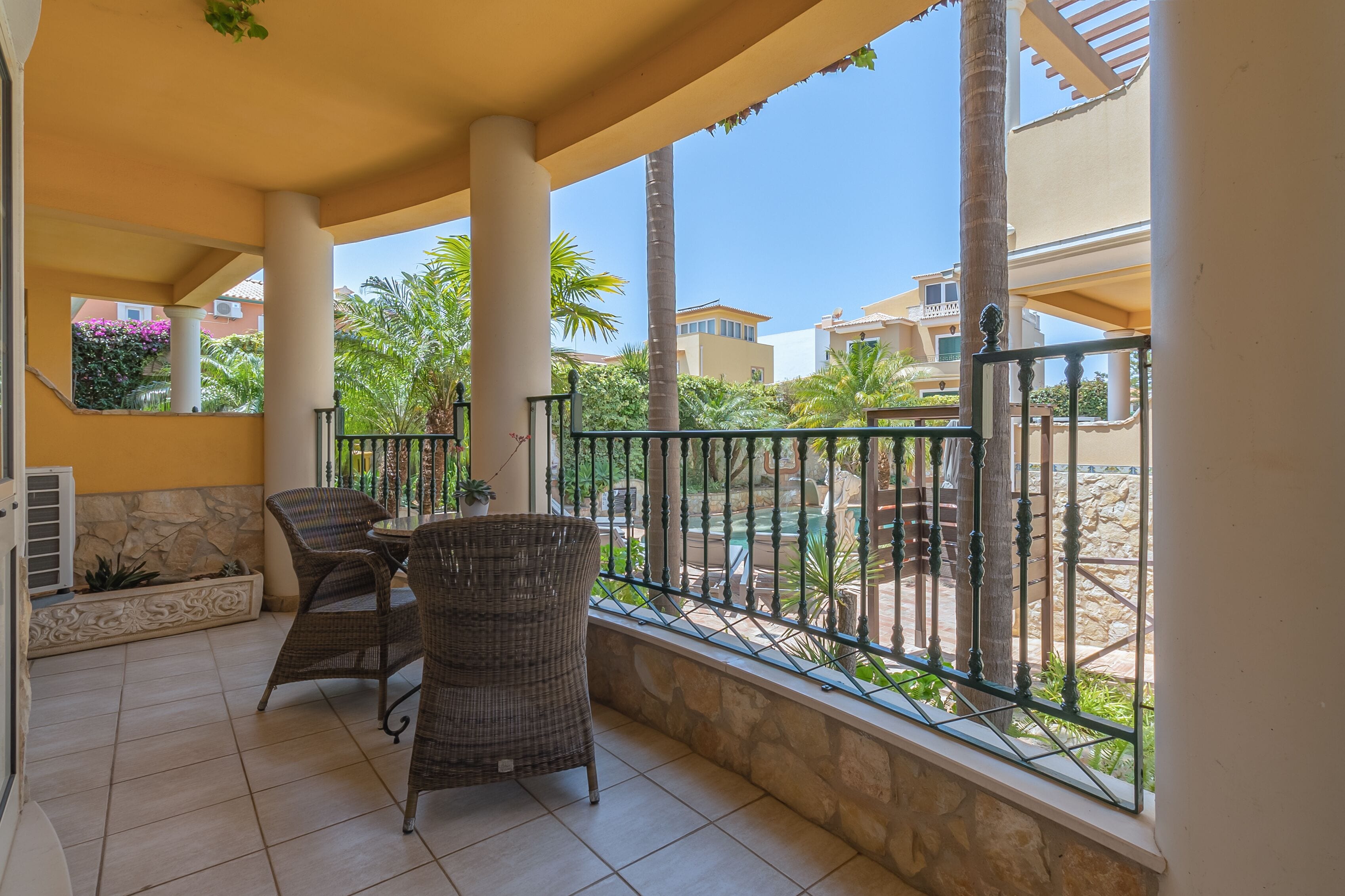 A balcony with wicker furniture, a stone planter, and a view of a pool and buildings.