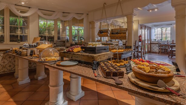 A buffet area with a variety of food items, including cakes and pastries, arranged on a counter with a tiled floor and large windows in the background. - Charming Residence & Guest House Dom Manuel I (Adults only) (Lagos)