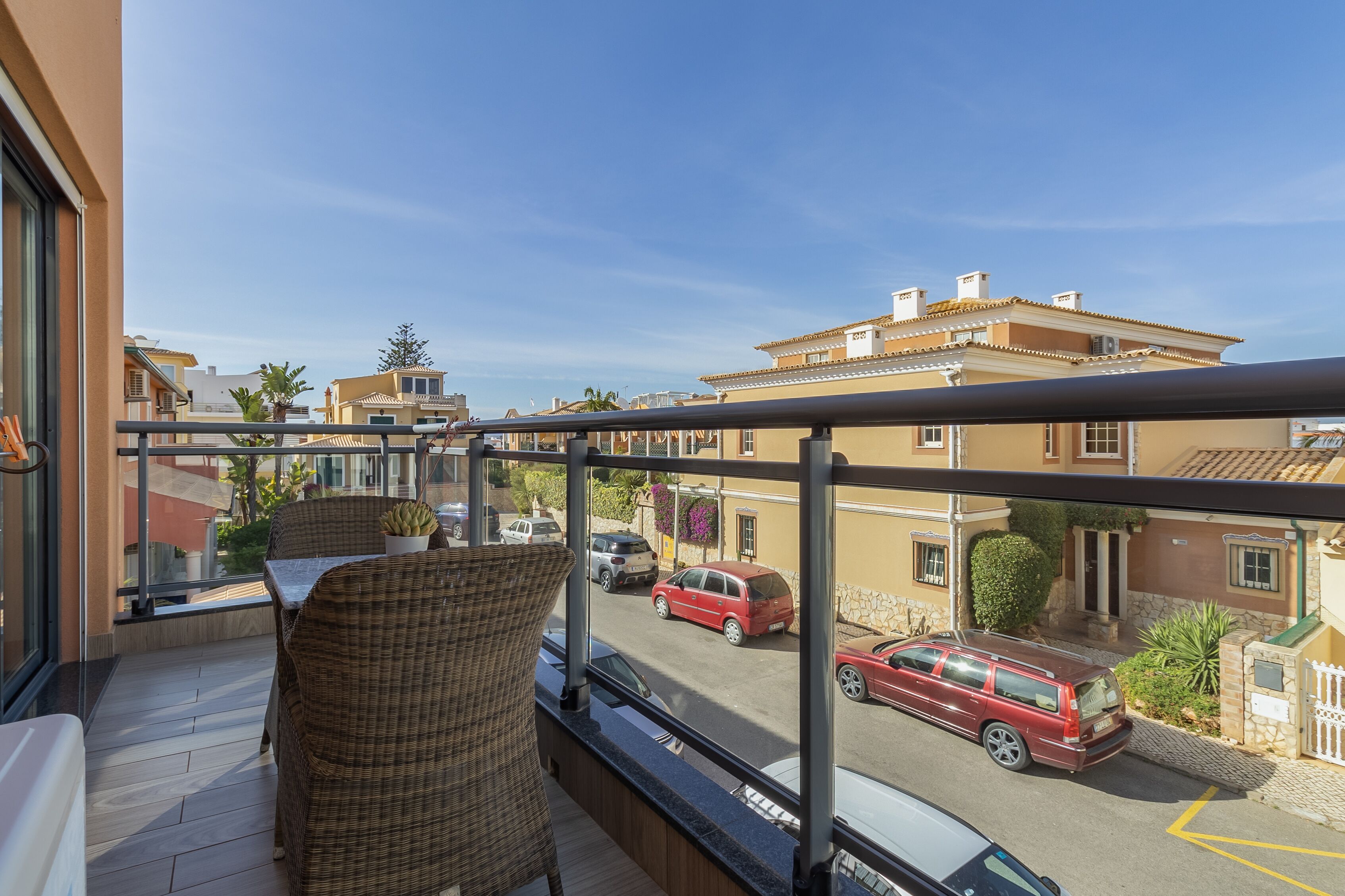 A balcony with wicker furniture, a view of a street with parked cars, and neighboring buildings.