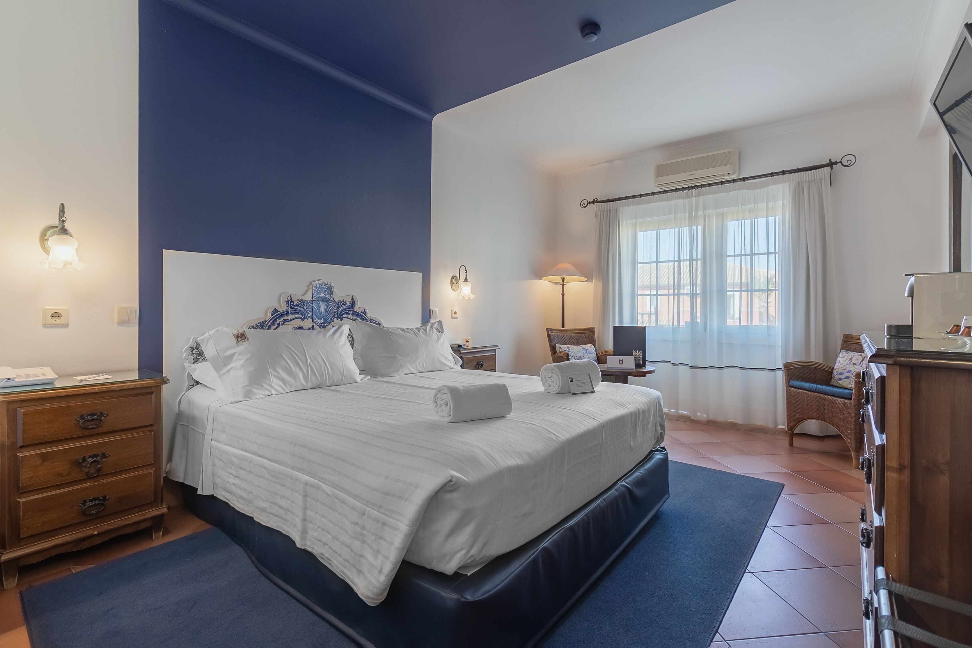Une chambre à coucher avec un grand lit, une commode, une table de chevet, une lampe et une fenêtre avec des rideaux.
