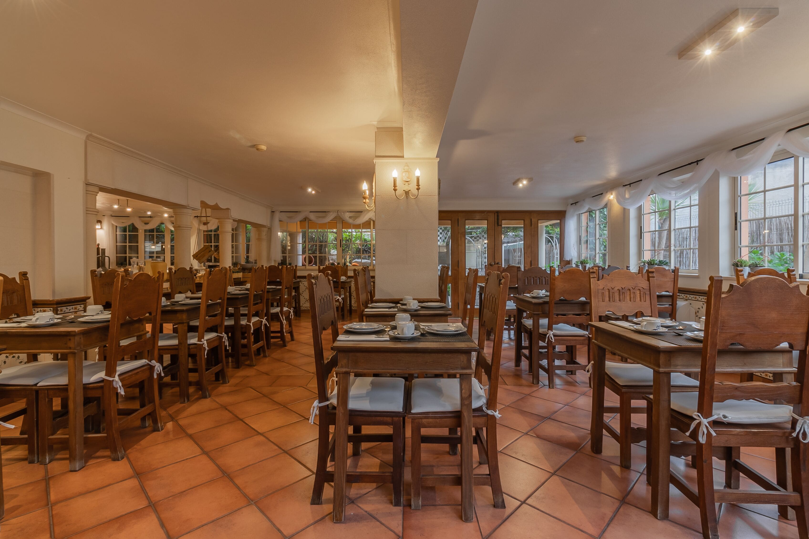 A spacious dining area with wooden tables and chairs, large windows, and a tiled floor.