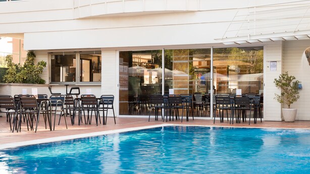 Zona de piscina con sillas y mesas, un edificio con grandes ventanales y un letrero que indica un restaurante.