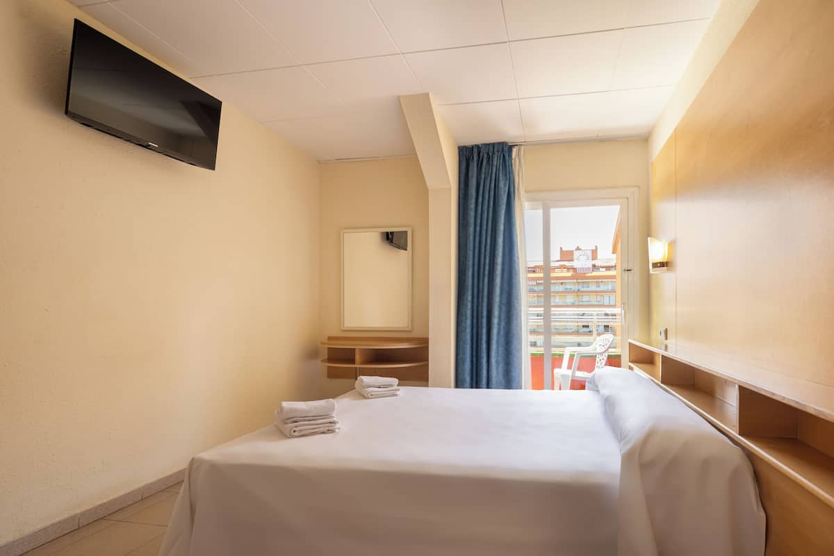 A hotel room with a bed, a flat-screen TV mounted on the wall, a mirror, a window with blue curtains, and a view of buildings outside.