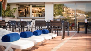 Fila de sillones de playa con toallas azules, un restaurante a la vista a través del ventanal y una zona de patio con baldosas.