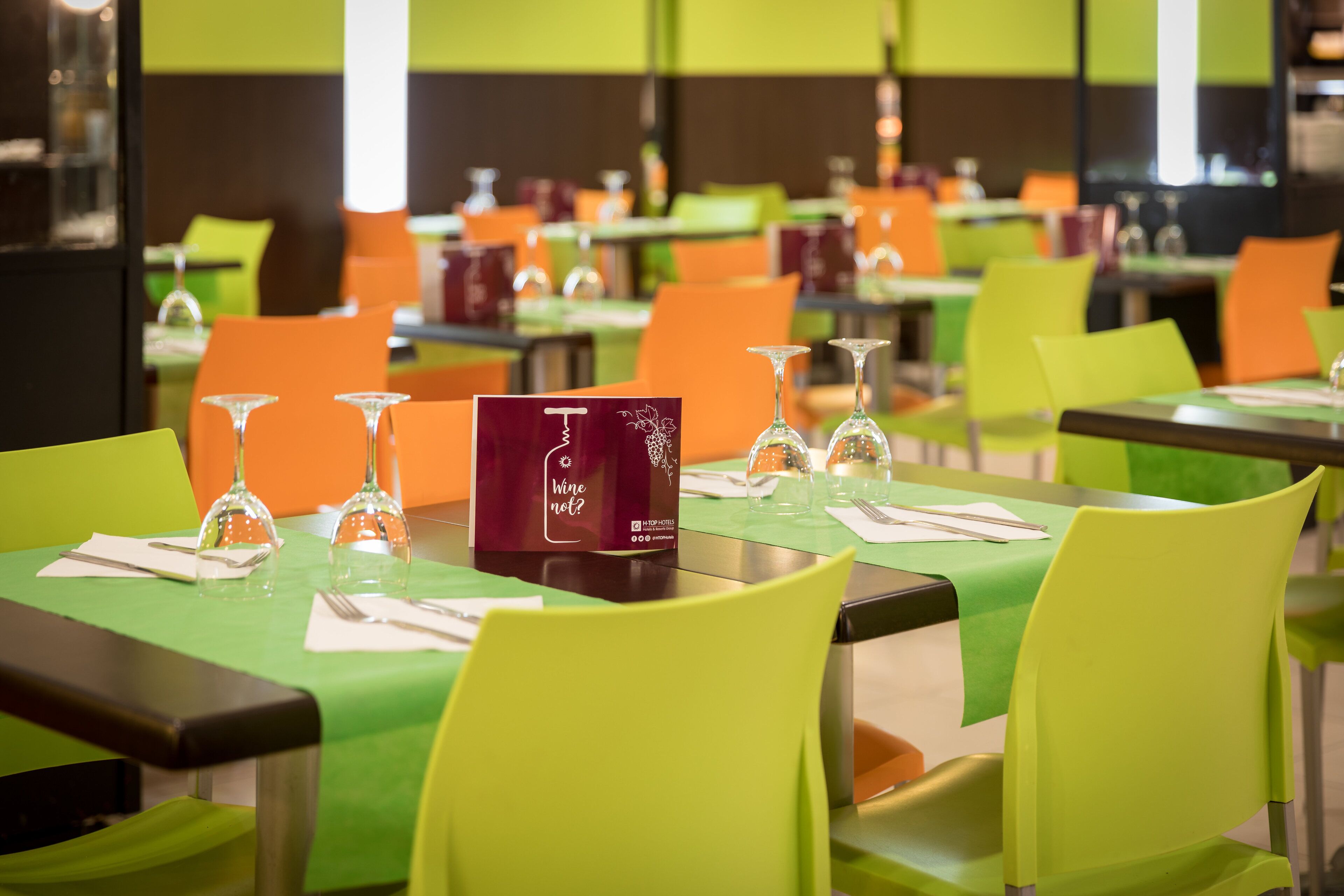 A restaurant with green tables and orange chairs, each set with wine glasses and menus.