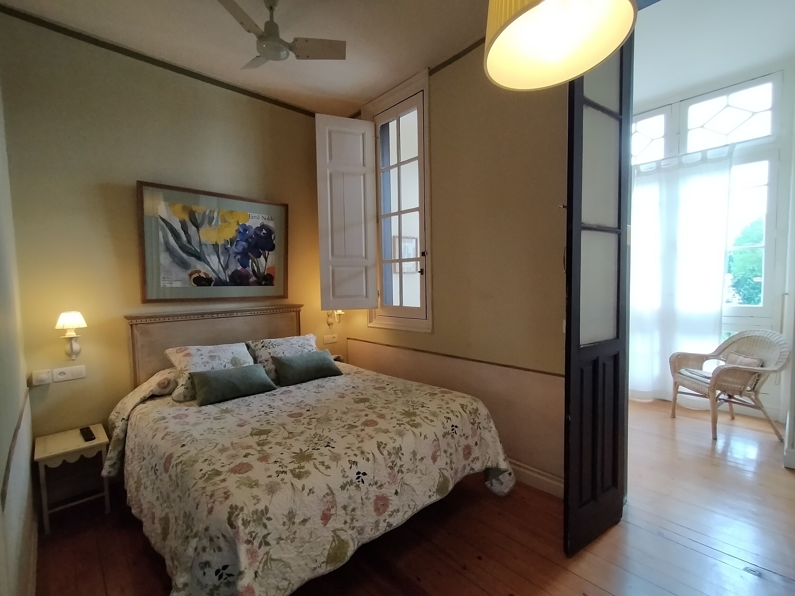 Un dormitorio con cama, mesita de noche, lámpara, ventilador de techo, ventana con contraventanas y una silla.