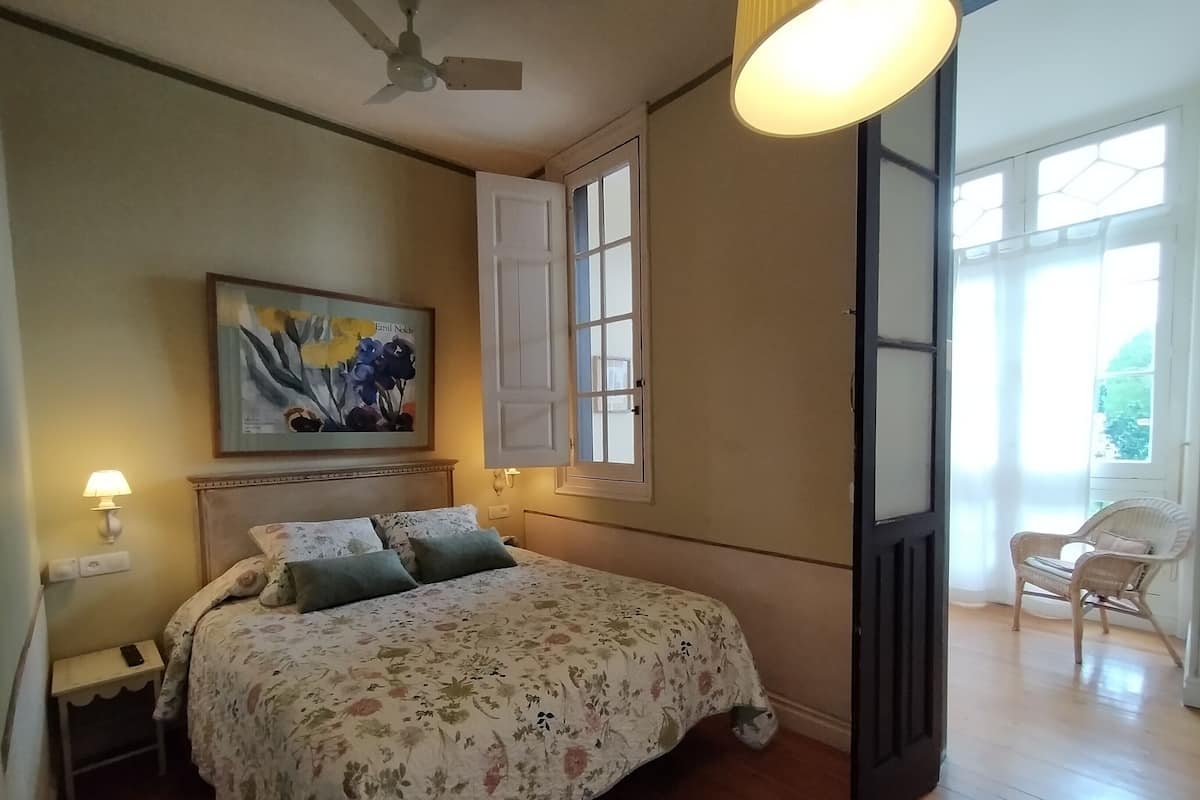 A bedroom with a bed, a nightstand, a lamp, a ceiling fan, a window with shutters, and a chair.