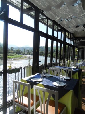 Un restaurant avec des tables dressées pour le service, de grandes fenêtres donnant sur une rivière et des places assises en plein air.