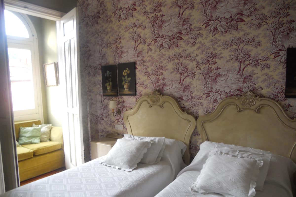 A bedroom with two beds, floral wallpaper, a window, and a sofa.
