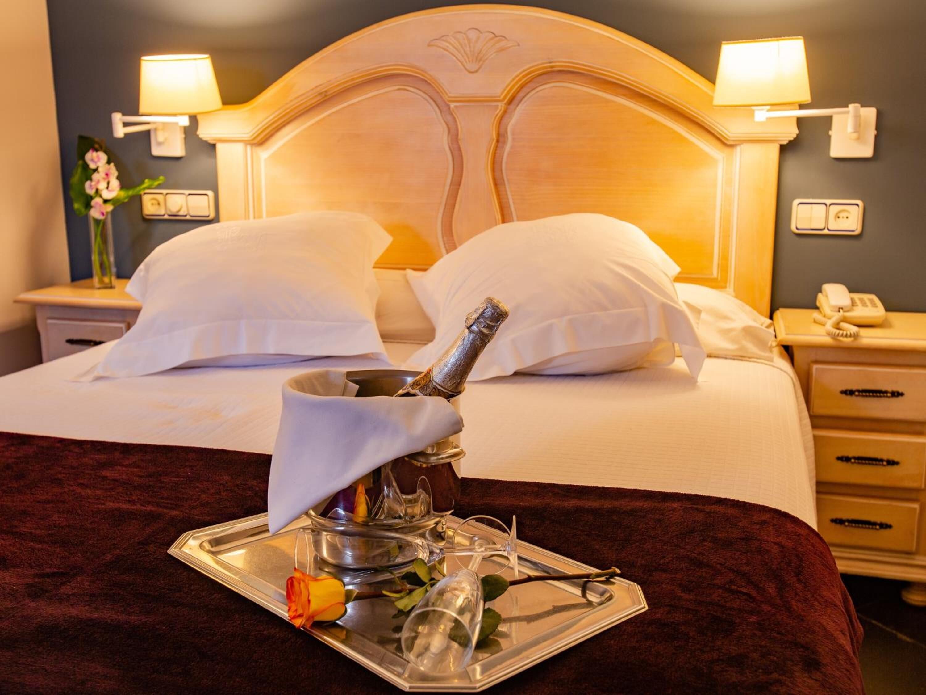 A hotel room with a bed, two bedside lamps, a nightstand with a phone, and a tray with a rose and a glass.