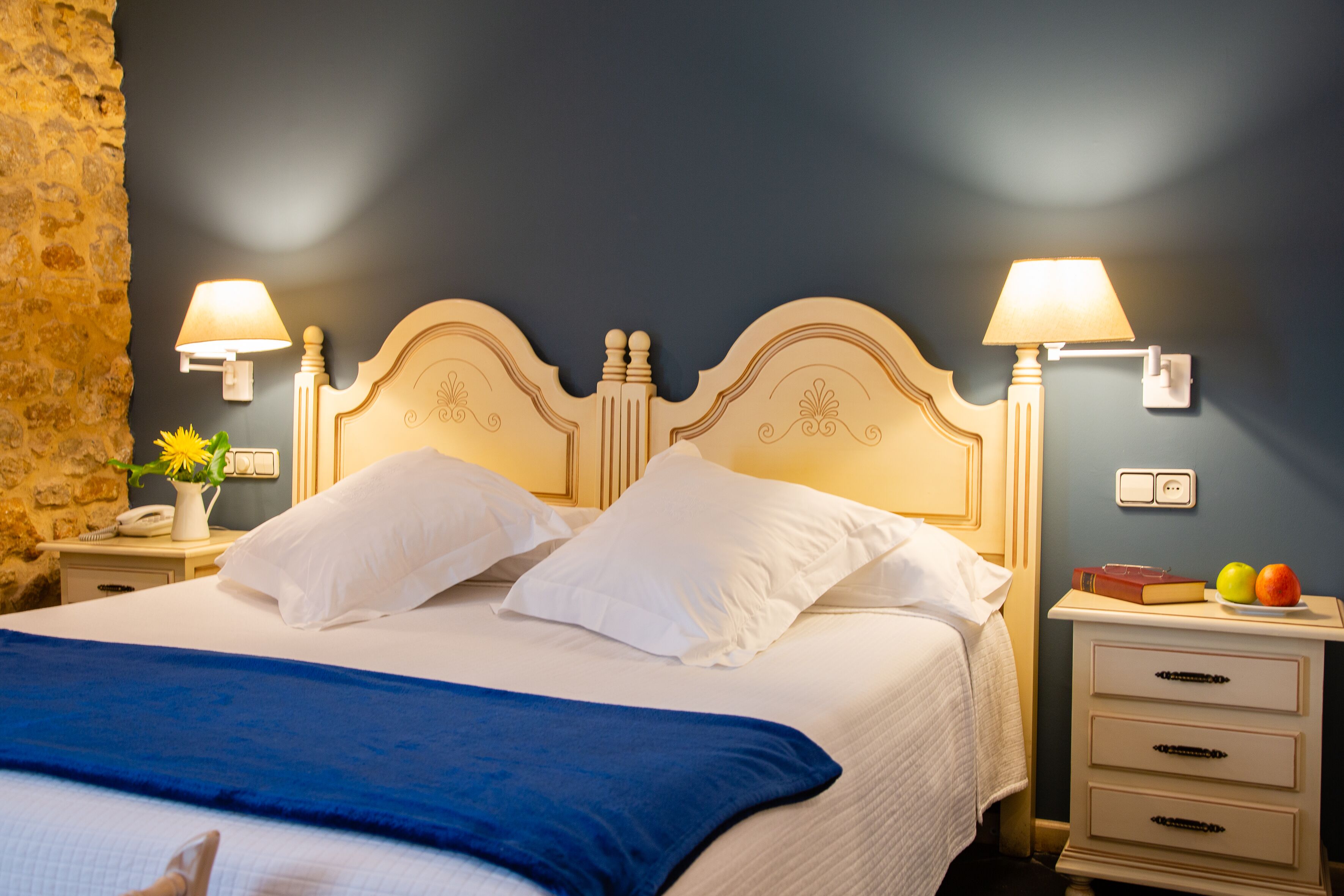 Una cama bien hecha con almohadas blancas y una manta azul, dos lámparas de noche, una mesita con un teléfono y un florero con flores, y un aparador con un libro y fruta.