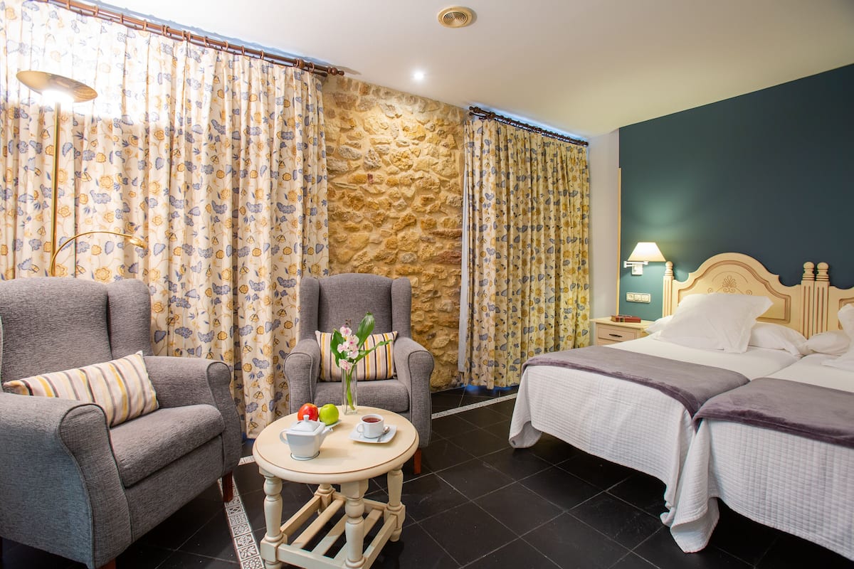 A hotel room with two single beds, a sitting area with a chair and table, a stone accent wall, and a nightstand with a lamp.