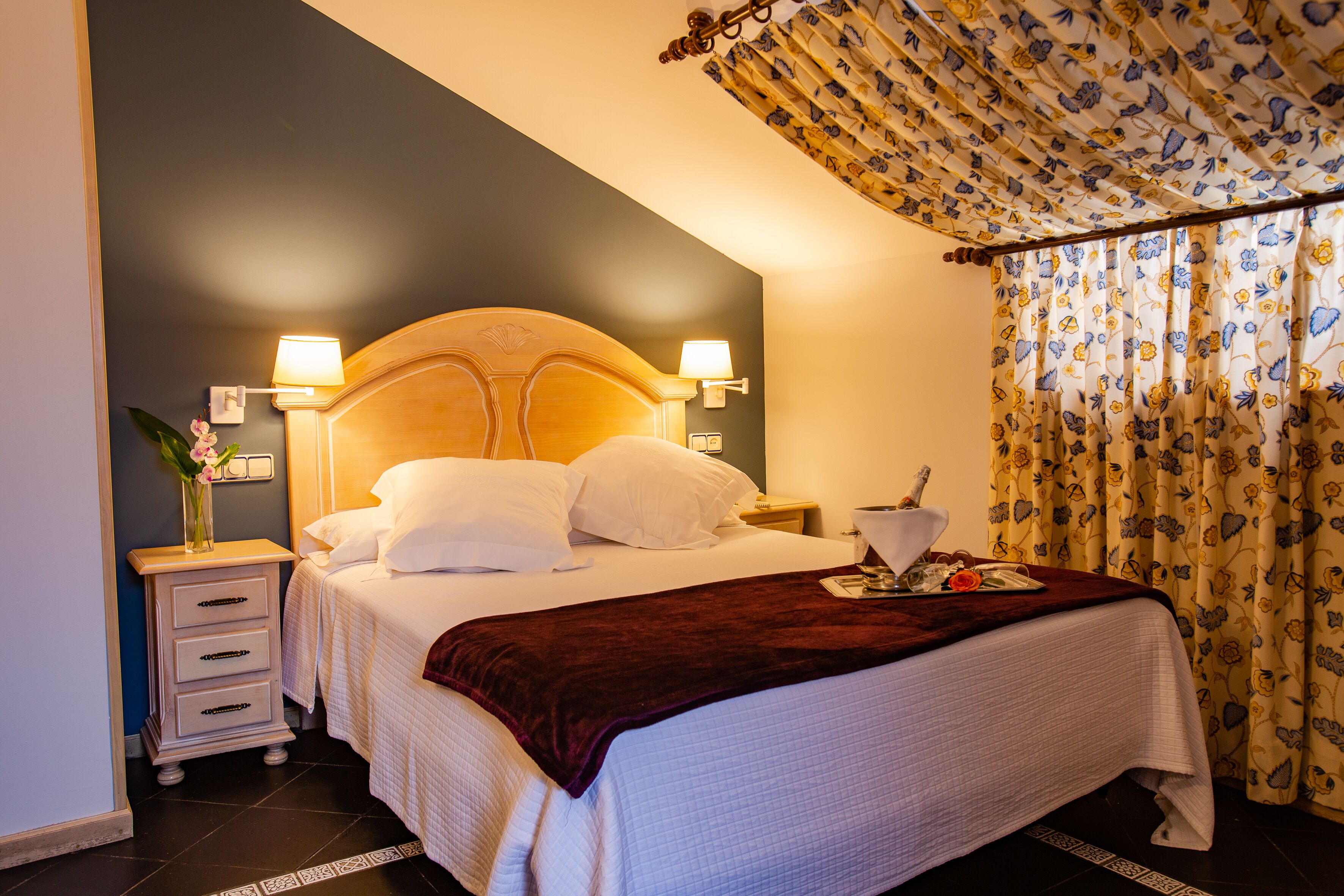 A neatly made bed with white linens and a dark red throw blanket, a bedside table with a lamp, and a floral curtain.