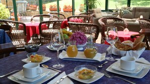 Mesa de jantar posta para o café da manhã, com frutas, pão e bebidas, em ambiente externo com cadeiras de vime e vista para o jardim.