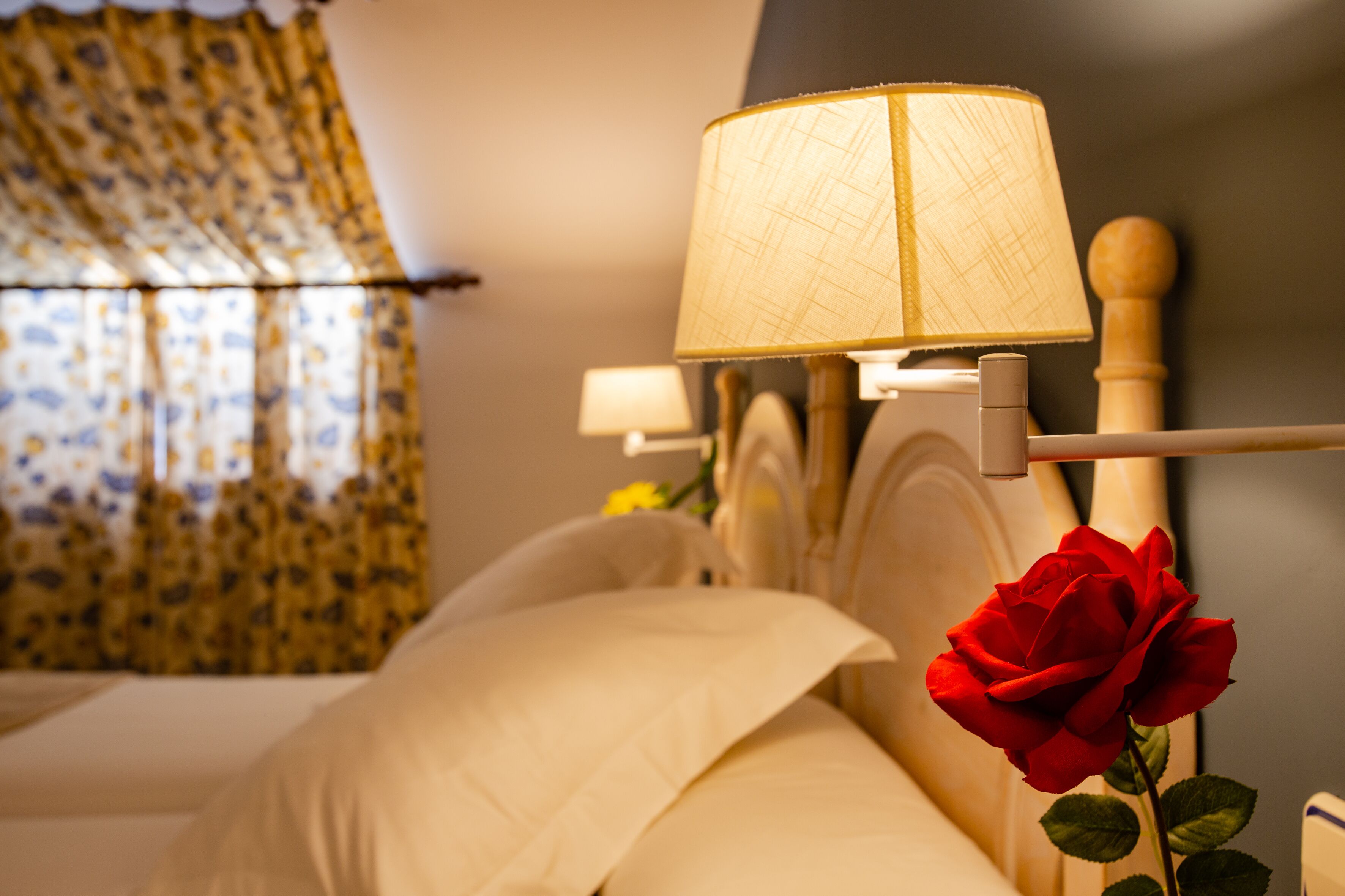 Un lit bien fait, avec du linge de lit blanc, une lampe de chevet et une rose rouge.
