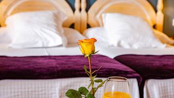 Un lit avec du linge de lit blanc et un plaid violet, une seule rose et un verre de jus d’orange sur la table de chevet.