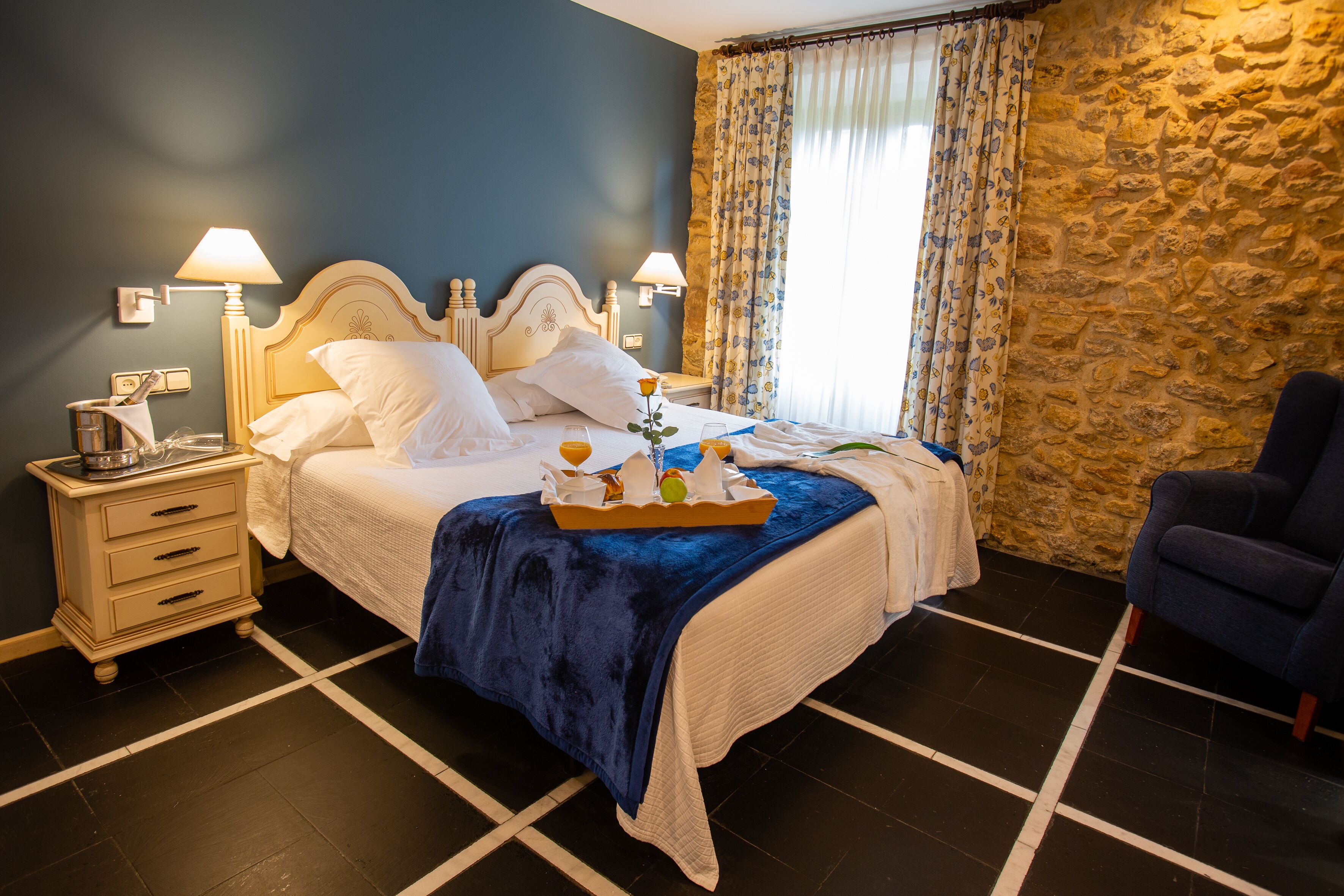 Una cama bien hecha con una manta azul, una mesita de noche con una lámpara y una silla en una habitación con paredes de piedra y un amplio ventanal con cortinas.