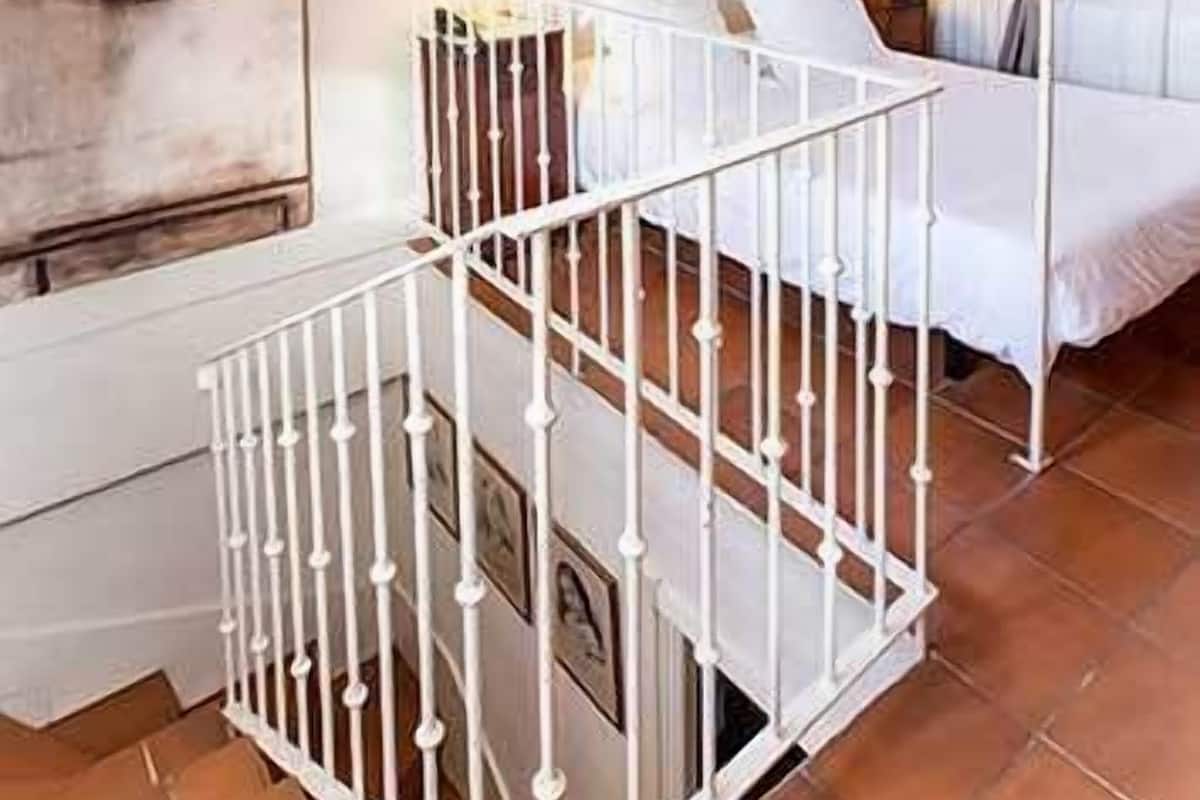 A room with a white metal crib, a bed with white bedding, a wall-mounted heater, and a window with curtains.