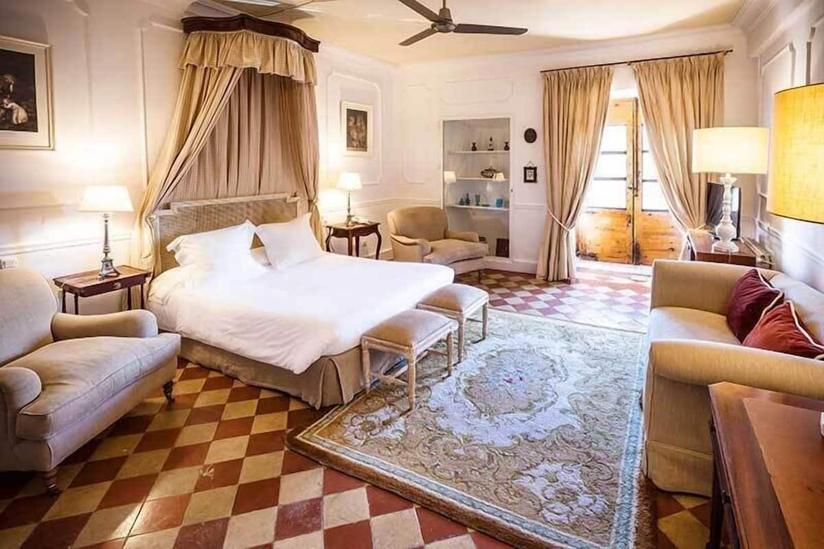 A hotel room with a large bed, two armchairs, a sofa, a small table, a bookshelf, and a large window with curtains.