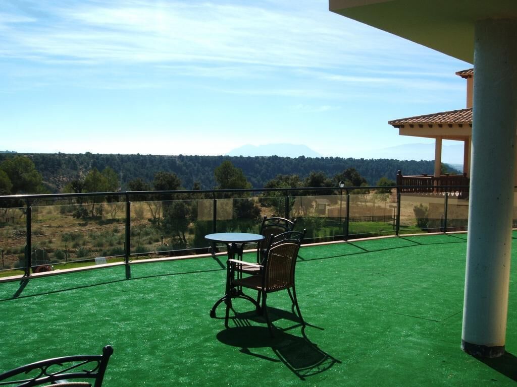 Un balcón con césped artificial verde, una mesa y dos sillas, y vistas a un paisaje boscoso.
