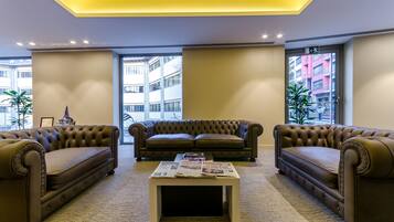 Un lobby moderno con un sofá de cuero marrón, una mesa de centro blanca con revistas y grandes ventanales que ofrecen vistas de la ciudad.
