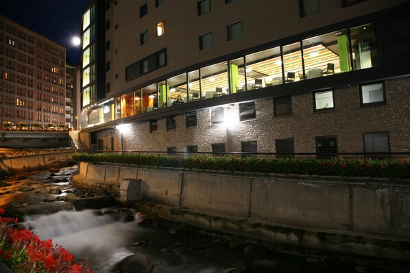 A modern building with a river in front, featuring a restaurant with a green interior and a view of the river.