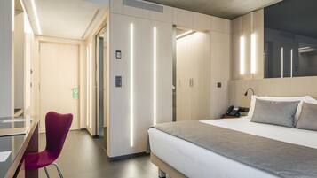 A modern hotel room with a large bed, a desk with a chair, and a wardrobe.