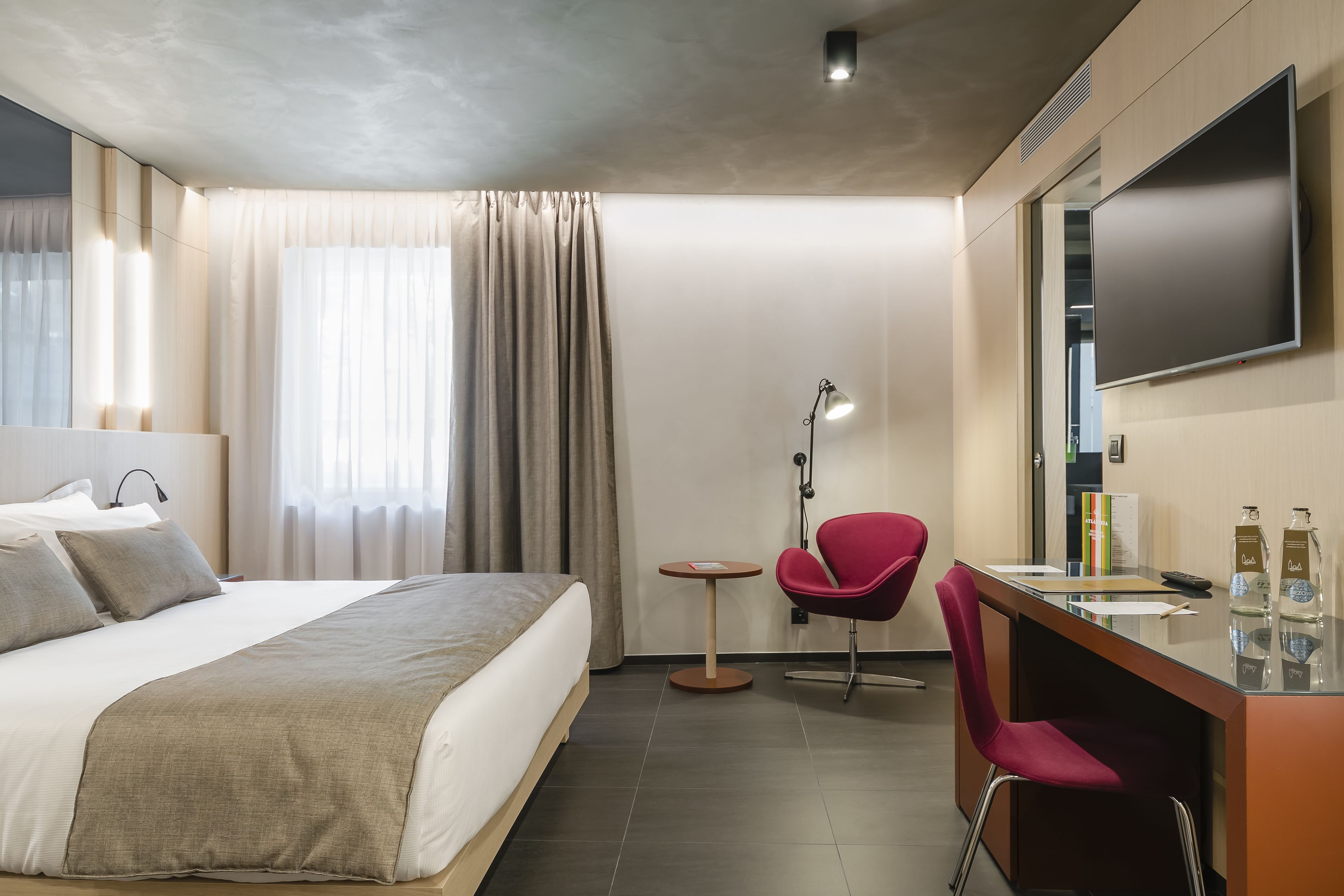 A hotel room with a bed, a desk, a chair, a TV, and a window with curtains.