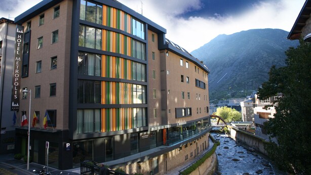 Un hotel moderno con fachada de vidrio, varios pisos y vistas a un río y montañas al fondo.